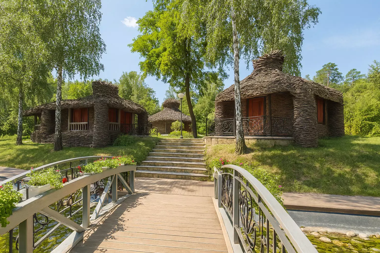 Un pod de lemn cu flori care duce spre căsuțe de basm făcute din crenguțe printre verdeață.